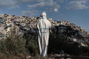 Wearing a protective suit, a worker stands before a towering mountain of waste, surveying the landfill's expansive waste and debris under a blue sky photo
