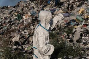 Dressed in a protective suit, a worker inspects a vast landfill, observing the layers of waste piled high and pointing out areas of concern for environmental impact photo