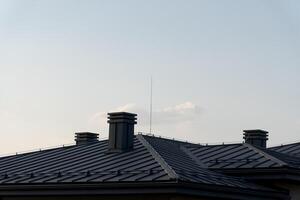 As the sun sets, a modern home's distinctive metal tile roof reflects soft hues of the sky. Tall chimneys add character while a delicate antenna reaches for the clouds, evoking tranquility photo