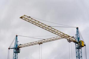 Two large construction cranes are actively positioned at a construction site, lifting materials as workers prepare the framework for a new house. The sky above is overcast photo