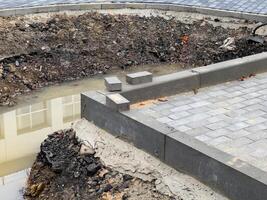 An unfinished brick pavement stretches alongside a sidewalk, featuring a trench filled with water. Loose soil and uninstalled pavers indicate ongoing construction activity photo