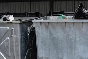 Two metal garbage containers filled with various types of rubbish are situated in a covered public space, collecting waste during daylight hours. The area shows signs of urban disarray photo