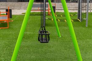 A bright green swing hangs motionless in a playground, inviting children to play while surrounded by lush grass and modern play equipment under a clear sky photo