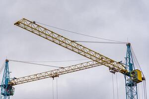 Two cranes are positioned at a construction site, with a yellow and blue structure against a backdrop of overcast skies, indicating active construction work in an urban environment photo