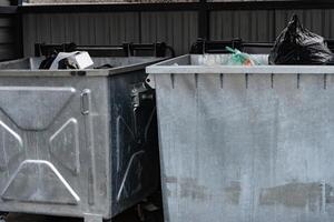 Two metallic garbage containers are positioned in a municipal area, overflowing with various types of waste, including bags and discarded items during daylight photo