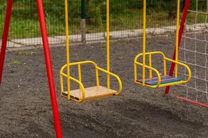 Two yellow swings with red supports stand still on a playground. The ground is made of gravel. There is a net fence behind the swings and green trees in the background photo
