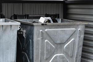 Two metallic garbage containers are positioned side by side, overflowing with various discarded items, captured during daylight in a city environment photo