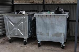 In an urban setting, two large metallic garbage containers are parked on wheels. One is filled with black trash bags, while the other contains various recyclable items photo
