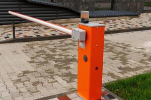 A bright orange barrier stands at a parking lot entrance, controlling vehicle access with its arm raised. The surrounding area features a paved surface and grassy edges photo