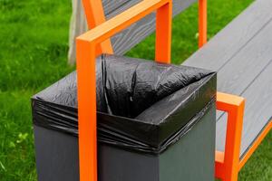 A black garbage container is attached to a park bench, showcasing a practical waste disposal solution in a well-kept grassy area, encouraging cleanliness in public spaces photo
