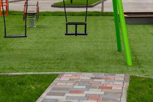 The playground features vibrant swings set on a soft green surface, surrounded by pathways, ready for children to explore, play, and have fun in the sun photo