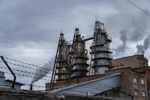 Thick smoke is released from the tall smokestacks of an industrial facility, indicating ongoing production. The sky is overcast, adding to the somber atmosphere photo