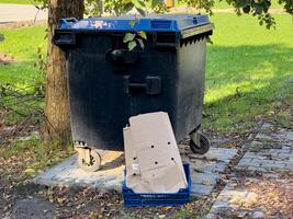 A black trash can stands next to a tree on the sidewalk, while a flattened cardboard box rests on the ground nearby, suggesting recent waste disposal in the area photo