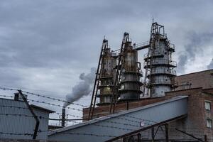 Thick smoke billows from the smokestacks of a manufacturing facility, contrasting against a cloudy sky. The industrial complex is surrounded by barbed wire fencing photo