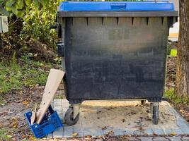 A black trash can sits on the street, surrounded by lush greenery. Beside it, a discarded cardboard piece and a blue crate are visible on the ground photo