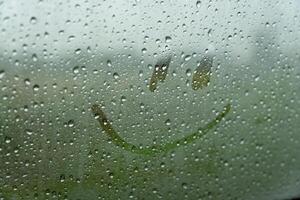 A simple smiley face is drawn on a foggy car window, surrounded by droplets of rain. The artwork brightens the dreary atmosphere on a rainy day photo