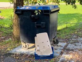 The black trash can sits near a tree on a street, with a cardboard box placed beside it, suggesting recent rubbish collection in a bright, outdoor environment photo