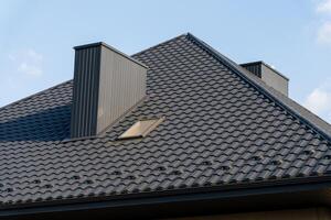 The roof of a contemporary house features sleek black metal material with distinct shingles, complemented by chimneys and a skylight, against a bright sky photo