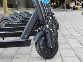 A row of electric scooters stands ready for riders along the busy city sidewalk, with pedestrians and vibrant surroundings adding to the lively atmosphere of the afternoon photo