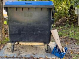 A black trash can with a blue lid sits on a sidewalk next to a cardboard box. The trash can is empty and the cardboard box is partially open. Concept of waste and neglect photo