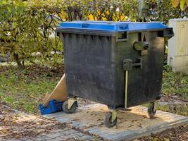 A sturdy black waste container is positioned next to a colorful autumn backdrop, surrounded by fallen leaves and debris. It's a sunny day, illuminating the scene photo