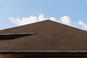 A brown shingle roof with a brown gutter against a blue sky with some white clouds photo