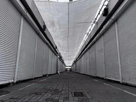 The narrow passageway of a once-busy market stands silent, lined with shuttered stalls. The overhead canopy casts shadows on the vacant ground, hinting at a forgotten vibrancy photo