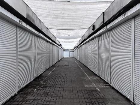 A deserted marketplace features rows of closed stalls with their shutters down, covered by a white canopy overhead, creating a quiet and still atmosphere photo