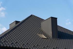 The structure features a sleek black tiled roof with two prominent chimneys rising, contrasted by a bright blue sky above, creating a contemporary aesthetic photo