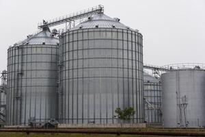 Several large metal silos are seen in a grain storage facility, surrounded by industrial structures, while a cloudy sky looms overhead photo