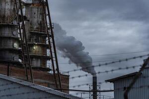 Smoke billows from smokestacks of an industrial facility, surrounded by buildings and fencing, against a backdrop of a moody, overcast evening sky photo