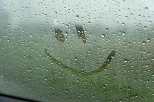 A single, simple smiley face is drawn on a car window. The window is covered in raindrops, obscuring the view outside. The smiley face is visible through the water droplets photo