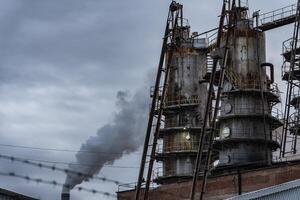 Tall, cylindrical structures with numerous metal platforms and ladders are situated against a gray sky with a thick plume of smoke rising from a nearby chimney photo