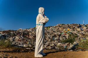 Dressed in protective clothing, an individual stands in front of a large landfill, reflecting on the environmental issues caused by waste accumulation under the clear blue sky photo