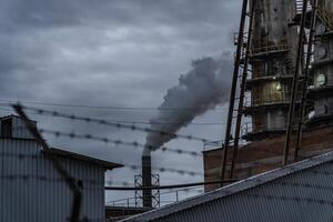 A large smokestack releases thick smoke into the air, surrounded by industrial buildings. The overcast sky adds a somber mood to the late afternoon scene photo