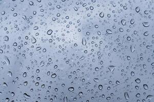 Raindrops cling to a glass surface, distorting the visibility of a gray sky. The droplets vary in size, reflecting light and contributing to the gloomy ambiance typical of rainy weather photo