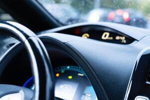 Raindrops on the windshield create a blurred backdrop while a driver's hand grips the steering wheel, focusing on the digital dashboard display photo