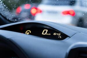 The dashboard shows zero kilometers per hour, indicating stopped traffic during rain, while cars are blurred in the background, creating a sense of delay photo