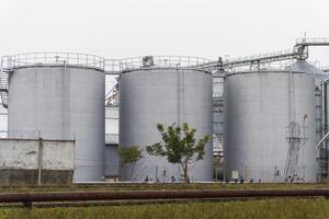 Three large, cylindrical storage tanks stand prominently at a grain processing facility, surrounded by an expansive agricultural landscape under cloudy skies photo