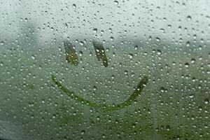 Raindrops create a textured surface on the glass, while a cheerful smiley face is drawn amidst the droplets, brightening the gloomy afternoon outside photo