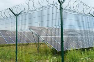 A security fence with barbed wire encloses a solar power farm featuring several solar panels tilted towards the sky. The grassy area has wildflowers and a cloudy backdrop photo