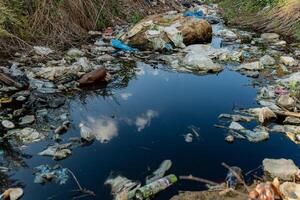 A riverbank is heavily littered with plastic waste and debris, showing environmental degradation. The still water reflects clouds, highlighting the contrast between nature and pollution photo