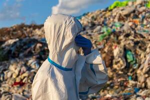 A person in a protective suit stands near a large landfill, covering their face while examining the piles of trash and debris under a sunny sky photo