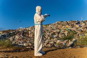 A person dressed in a protective suit stands at a landfill, looking out over heaps of waste, surrounded by blue skies and scattered debris, emphasizing environmental concerns photo