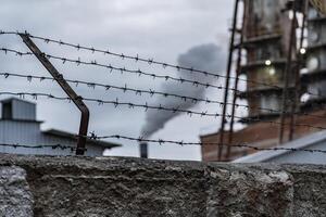 A barbed wire fence stands in the foreground, with a factory in the background exhaling smoke into the gray sky, reflecting an industrial environment photo