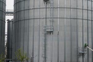 Tall metallic silos rise against the cloudy sky, showcasing access ladders and surrounding agricultural land. The structures store grains, vital for farming photo