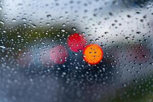 Raindrops create a textured pattern on a car window, blurring red and orange traffic lights in the background during a rainy day, revealing a typical urban scene photo