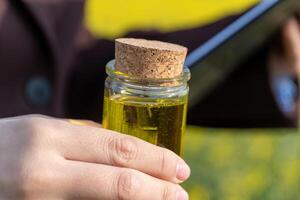 Hands hold clear glass bottle with cork stopper filled with golden liquid over blurred yellow field photo