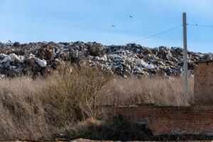 Massive Garbage Dump with Debris and Waste Creating a Hill Under Blue Sky near Red Brick Wall and Dry Grass photo