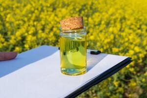 A hand holds a small glass jar filled with biodiesel near a clipboard, set against a vibrant field of yellow flowers. Quality testing is underway in biodiesel research photo